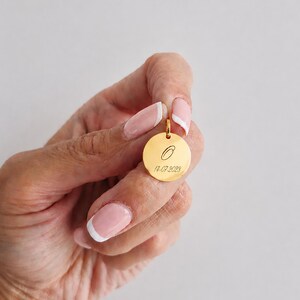 Puede incluir: Un colgante dorado con dise&ntilde;o circular. El colgante presenta la letra "O" y la fecha "14-07-2023" grabadas en su superficie. Cuelga de un peque&ntilde;o lazo en la parte superior.
