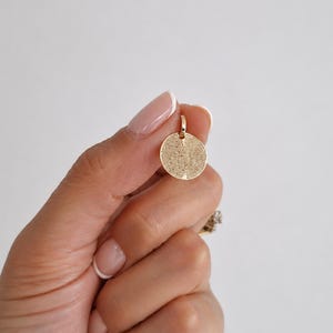 Puede incluir: Un colgante circular dorado con un dise&ntilde;o de huella dactilar. El colgante est&aacute; suspendido de un peque&ntilde;o lazo en la parte superior. El patr&oacute;n de huellas dactilares cubre toda la superficie del colgante.