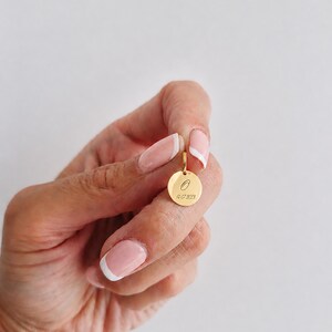 Puede incluir: Un colgante dorado con un dije circular. El dije presenta una letra "O" grabada y la fecha "14-07-2023". El colgante se muestra sobre un fondo blanco.