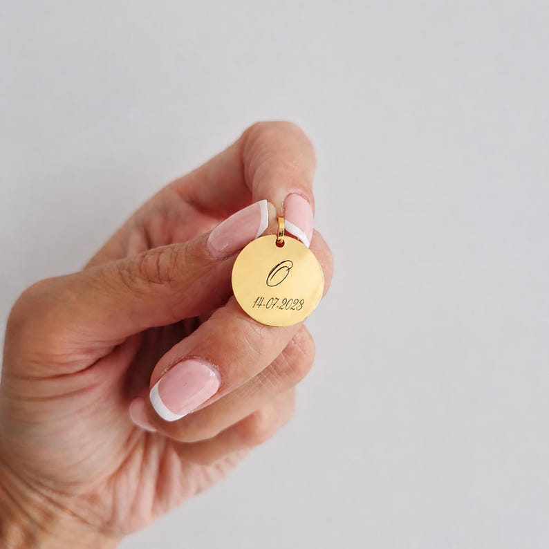 Puede incluir: Un colgante redondo dorado con un peque&ntilde;o lazo en la parte superior. El colgante tiene una letra "O" grabada y la fecha "14-07-2023". El colgante se sostiene en una mano con u&ntilde;as cuidadas.