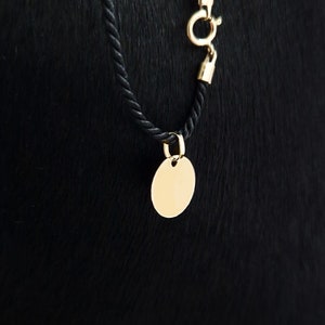 Puede incluir: Un colgante ovalado dorado cuelga de un collar de cuerda negro. El colgante es liso y en blanco, con un peque&ntilde;o agujero en la parte superior para la fijaci&oacute;n. El collar tiene un cierre dorado.