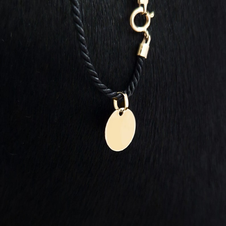 Puede incluir: Un colgante ovalado dorado cuelga de un collar de cuerda negro. El colgante es liso y en blanco, con un peque&ntilde;o agujero en la parte superior para la fijaci&oacute;n. El collar tiene un cierre dorado.