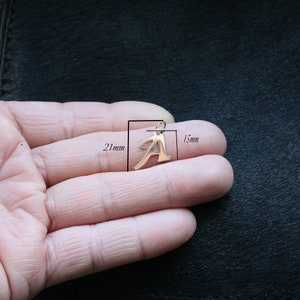 Puede incluir: Un colgante de metal en tono dorado con la forma de la letra may&uacute;scula A. El colgante presenta una peque&ntilde;a anilla en la parte superior para su sujeci&oacute;n. Las anotaciones t&eacute;cnicas indican una altura de 21 mm y una anchura de 15 mm de escala.