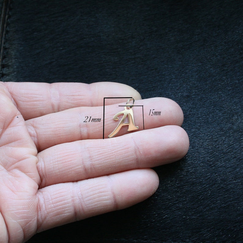 Puede incluir: Un colgante de metal en tono dorado con la forma de la letra may&uacute;scula A. El colgante presenta una peque&ntilde;a anilla en la parte superior para su sujeci&oacute;n. Las anotaciones t&eacute;cnicas indican una altura de 21 mm y una anchura de 15 mm de escala.