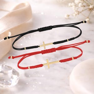 May include: Two adjustable bracelets with gold cross pendants. One bracelet is black, the other is red. Both have gold accents and a sliding knot closure. The bracelets are displayed on a light background with soft lighting.