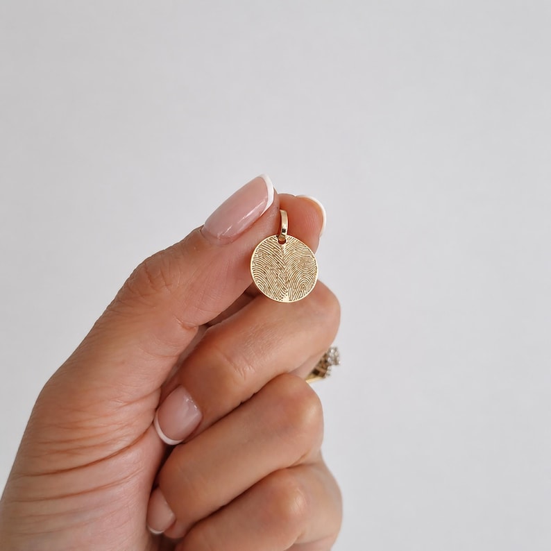 Puede incluir: Un colgante circular dorado con un dise&ntilde;o de huella dactilar. El colgante est&aacute; sostenido por una mano sobre un fondo blanco. El colgante mide aproximadamente 2 cm de di&aacute;metro y tiene un peque&ntilde;o lazo en la parte superior para una cadena.