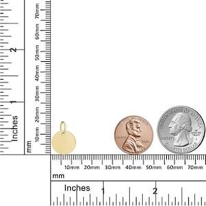 Puede incluir: Un colgante circular dorado con un peque&ntilde;o lazo para una cadena. El colgante se muestra junto a una regla, un centavo y un cuarto de d&oacute;lar para comparar el tama&ntilde;o. El colgante tiene aproximadamente 1,3 cm de di&aacute;metro.