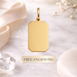 May include: A gold-coloured rectangular pendant with a loop at the top for a chain. The pendant has a smooth surface and is displayed with a white ribbon and a sign that reads "FREE ENGRAVING".