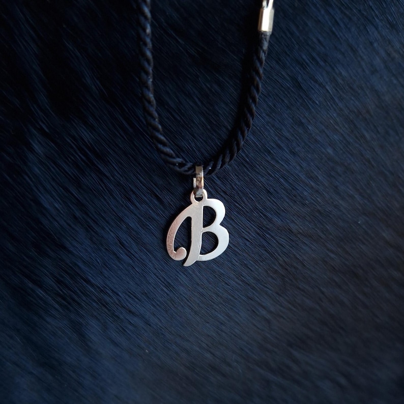 Puede incluir: Un colgante de metal plateado con forma de letra B may&uacute;scula, suspendido de un collar de cord&oacute;n trenzado negro. El colgante presenta un acabado liso y pulido y est&aacute; sujeto al cord&oacute;n mediante una peque&ntilde;a y sencilla anilla de metal plateado.