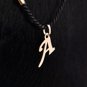 Puede incluir: Un colgante de metal de color dorado con forma de letra may&uacute;scula A, con una tipograf&iacute;a de estilo manuscrito. El colgante est&aacute; unido a una peque&ntilde;a anilla circular, suspendida de un collar de cord&oacute;n trenzado negro con un terminal met&aacute;lico dorado.