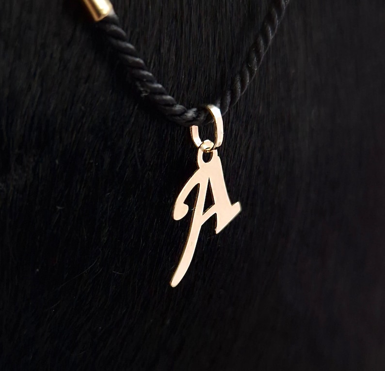 Puede incluir: Un colgante de metal de color dorado con forma de letra may&uacute;scula A, con una tipograf&iacute;a de estilo manuscrito. El colgante est&aacute; unido a una peque&ntilde;a anilla circular, suspendida de un collar de cord&oacute;n trenzado negro con un terminal met&aacute;lico dorado.