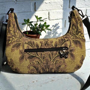 Może przedstawiać: Półksiężycowa torebka z kwiatowym wzorem w odcieniach brązu i złota. Torebka posiada czarny zamek błyskawiczny, zawieszkę w kształcie motyla oraz czarny, regulowany pasek na ramię. Mała roślina w brązowej doniczce znajduje się za torebką.