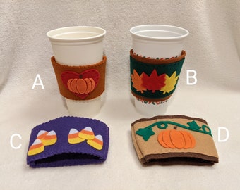 May include: Four felt coffee cup sleeves with fall-themed designs. One sleeve features a pumpkin inside a heart, another has colorful leaves, a third has candy corn, and the last has a pumpkin with vines. The sleeves are designed to fit around standard coffee cups.