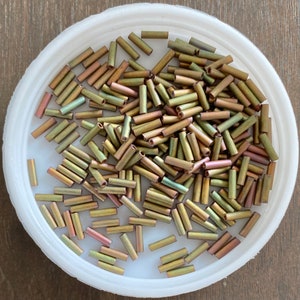 May include: A white bowl filled with a large quantity of small, gold-toned, glass beads. The beads are cylindrical in shape and have a slightly iridescent finish.