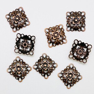 May include: Eight antique bronze-coloured metal charms with a square shape and intricate floral designs. Each charm features a raised flower in the centre with delicate filigree details.