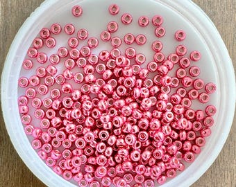 Puede incluir: Un recipiente blanco lleno de cuentas metálicas de color rosa. Las cuentas son pequeñas y redondas, y tienen un acabado brillante.