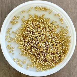 May include: A white, circular container filled with numerous small, gold-coloured beads. The beads are closely packed, filling the container. The beads appear cylindrical and have a metallic sheen. The container rests on a wooden surface.