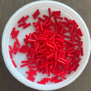 May include: A bowl filled with red glass beads. The beads are cylindrical in shape and are all the same size.