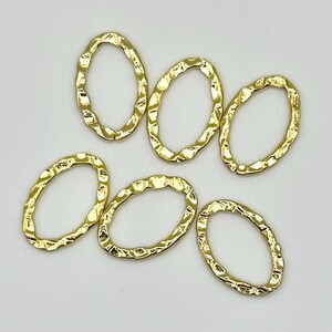 May include: Six gold-coloured oval charms with a hammered texture are scattered on a white surface. The charms are open ovals, and the surface of each charm has a textured, irregular pattern. The charms are likely for jewellery making.