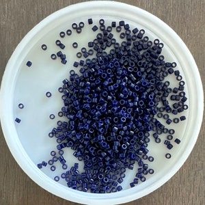May include: A white bowl filled with dark blue seed beads. The beads are small and round, and they are arranged in a random pattern.