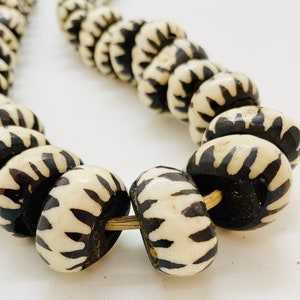 May include: A close-up of a necklace made of black and white beads. The beads are round and have a pattern of white stripes on a black background. The necklace is strung on a gold cord.