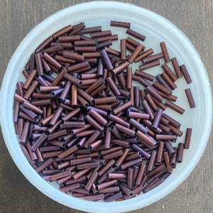 May include: A bowl containing around 100 small, brown, metallic, cylindrical beads. The beads are approximately 1.2 cm long and have a slightly iridescent finish.