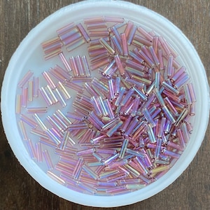 May include: A white bowl filled with iridescent pink glass beads. The beads are long and cylindrical, with a slightly rounded shape.