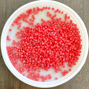 May include: A white plastic container filled with numerous small, coral-coloured, cylindrical beads. The beads are scattered across the container, creating a textured surface. The container sits on a wooden surface.