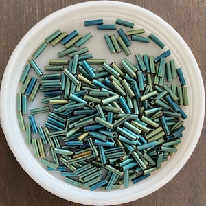 May include: A bowl filled with teal and gold glass beads. The beads are long and cylindrical, and they are arranged in a random pattern.