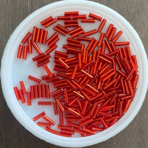 May include: A bowl filled with orange glass beads. The beads are long and thin, and they are arranged in a random pattern.