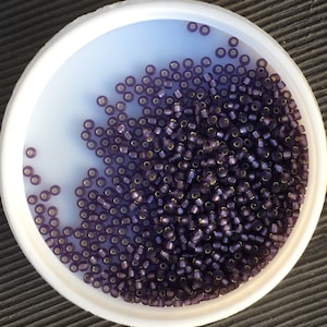 May include: A white plastic container filled with small, dark purple seed beads. The beads have a metallic gold-coloured hole. The container is on a dark surface. These beads are ideal for jewellery making and crafts.