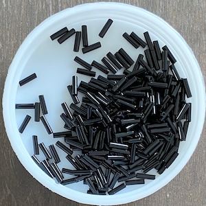 May include: A white bowl filled with black glass beads. The beads are long and thin, resembling small cylinders.