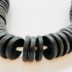 May include: Close-up of a dark brown wooden necklace. The necklace is made of circular, flat wooden beads strung together on a white cord. The beads have a slightly rough texture and are evenly spaced.