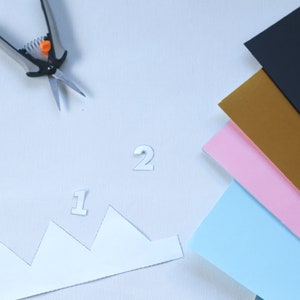 Puede incluir: Una corona de papel blanco con los n&uacute;meros "1" y "2" en ella. Tambi&eacute;n hay tijeras, una hoja de papel negro, una hoja de papel dorado, una hoja de papel rosa y una hoja de papel azul claro.