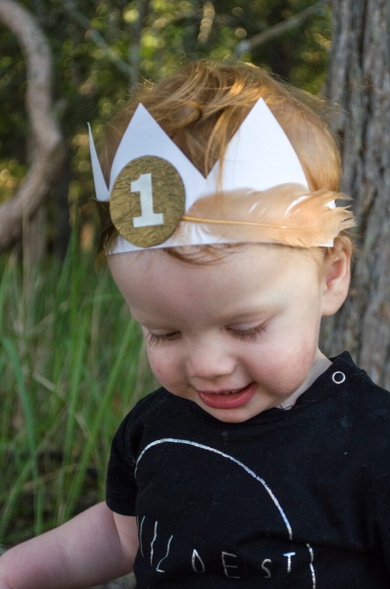 Puede incluir: Un ni&ntilde;o peque&ntilde;o con una corona de papel blanco con un c&iacute;rculo dorado y el n&uacute;mero "1" en ella. La corona tiene una pluma marr&oacute;n unida a ella. El ni&ntilde;o lleva una camiseta negra con la palabra "WILDEST" impresa en ella.