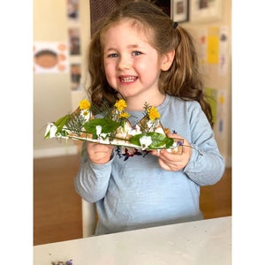 Puede incluir: Una ni&ntilde;a sostiene una corona de cart&oacute;n decorada con hojas verdes, flores blancas y flores amarillas.