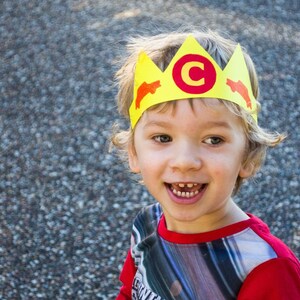 Puede incluir: Un ni&ntilde;o peque&ntilde;o con una corona de papel amarilla con una C roja y dise&ntilde;os de peces naranjas. El ni&ntilde;o sonr&iacute;e y lleva una camisa a rayas rojas y grises.