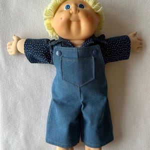 Peut inclure: Une poupée à la peau claire, aux yeux bleus et aux cheveux courts en laine jaune. La poupée porte une chemise bleu foncé avec un motif à petits points blancs et orange sous une salopette en jean bleu avec deux boutons bleus.