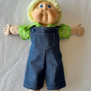 May include: A doll with light skin, blue eyes, and short yellow yarn hair. The doll wears a green short-sleeved shirt with a small white circular pattern and dark blue denim overalls with two dark blue buttons at the top of the bib.