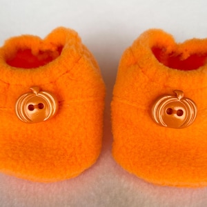May include: A pair of orange fleece baby booties, each with a pumpkin-shaped button. The booties are a vibrant orange and have a soft, fluffy texture. The buttons are the same orange colour and provide a decorative element. Ideal for a baby's Halloween outfit.