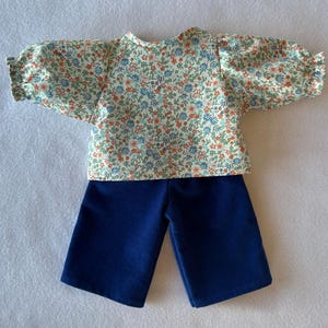 May include: A two-piece outfit featuring a floral print top and navy blue shorts. The top has short, puffed sleeves and a cream background with small orange, blue, and green flowers. The shorts are a solid, dark blue color.