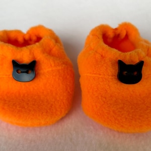 May include: A pair of bright orange fleece baby booties, each adorned with a black cat face button. The booties have a soft, fluffy texture and are a vibrant orange. The black cat buttons provide a contrasting detail.