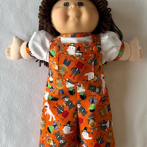 May include: A doll with brown yarn hair and a round face wearing a white short-sleeved shirt under orange overalls. The overalls feature a repeating pattern of Halloween characters including ghosts, devils, mummies, and pumpkins on a bright orange fabric.