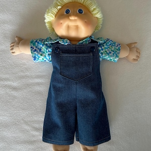 May include: A doll with light skin, blue eyes, and blonde yarn hair. The doll wears a short-sleeved shirt with a blue, green, and white pixelated pattern underneath dark blue denim overalls featuring a front pocket and two dark buttons.