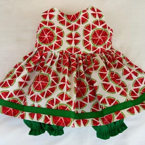 May include: A sleeveless dress featuring a white background with a repeating red and green watermelon slice pattern. The dress has a gathered skirt with a horizontal green ribbon trim near the hem and is paired with solid green ruffled bloomers.