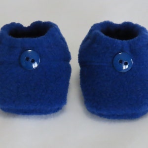 Peut inclure: Une paire de chaussons de bébé bleu roi en polaire douce. Chaque chausson est orné d'un bouton bleu assorti pour la fermeture. Les chaussons sont conçus pour garder les pieds de bébé au chaud.