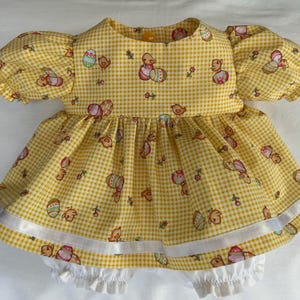 May include: A yellow gingham baby dress and bloomers set. The dress has short, puffed sleeves and is patterned with Easter eggs and chicks. White ribbon trim accents the hem. The bloomers are white with ruffled edges. A charming outfit.