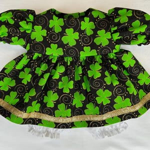 May include: A baby dress in green and black with a clover design. The dress has short, puffed sleeves and a gold trim. The fabric displays green clovers, gold swirls, and dots on a black backdrop.