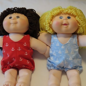May include: Two Cabbage Patch Kids dolls wearing summer outfits. One doll has brown curly hair and is wearing a red swimsuit with white anchors. The other doll has yellow curly hair and is wearing a blue swimsuit with white sailboats.