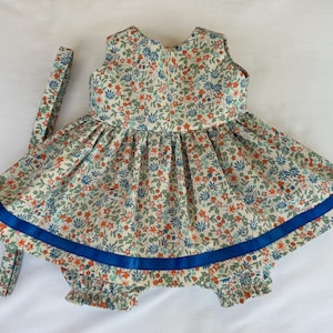 Peut inclure: Une robe sans manches et une culotte assortie avec un fond crème et un motif floral dense composé de petites fleurs bleues, orange et vertes. La robe présente une jupe froncée avec un ruban bleu roi horizontal près de l'ourlet.